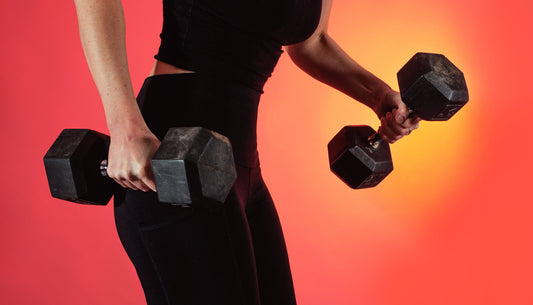 A person dressed in black holds two black weights and lifts one up. They are standing in front of a yellow and red background.